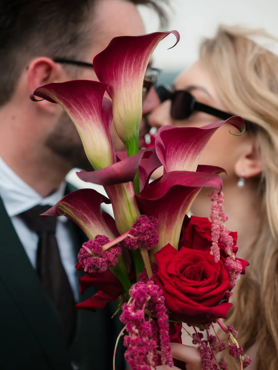 A bouquet of deep red callas and roses in the foreground while the couple kiss behind it