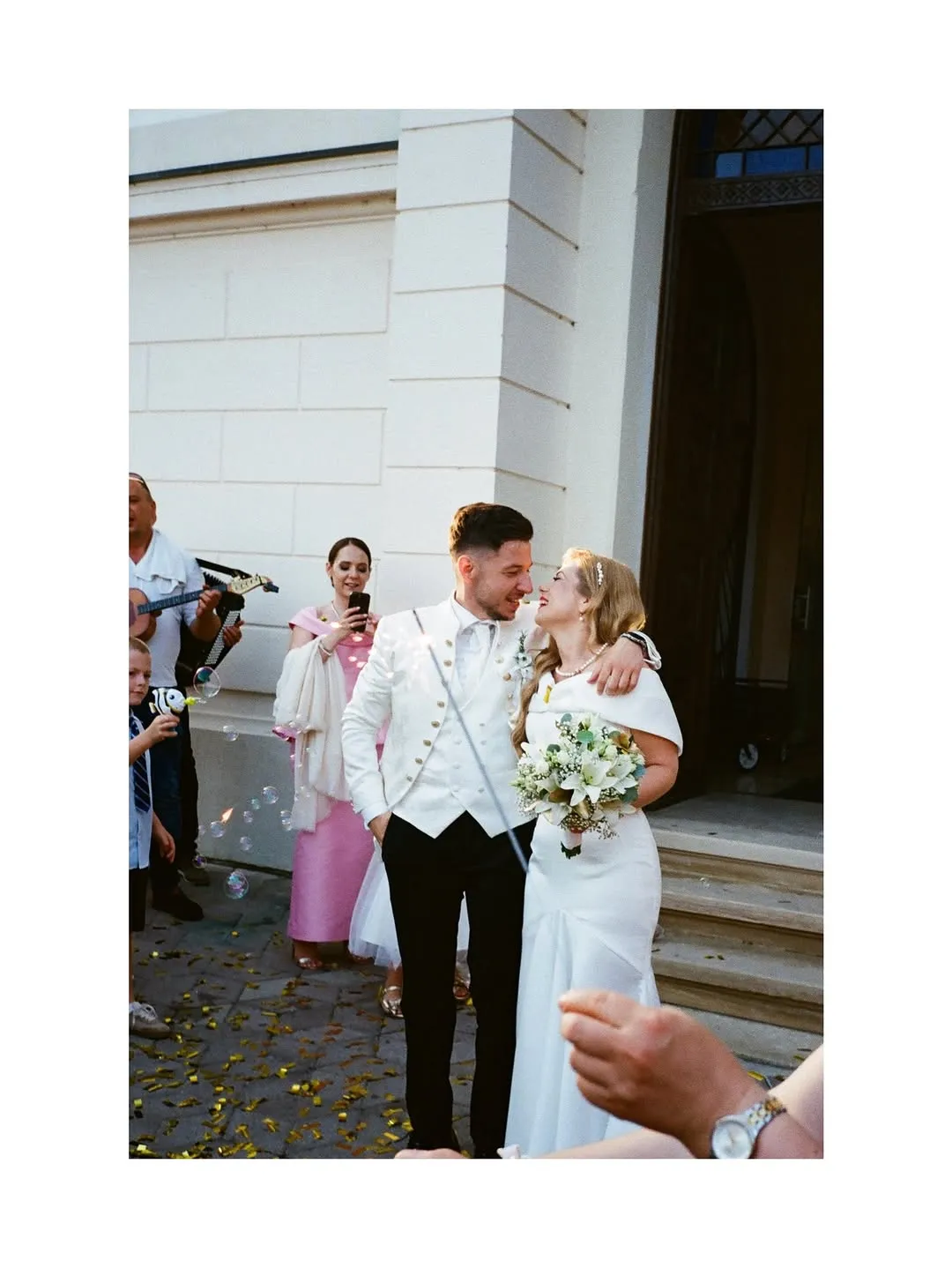 Guests greet the smiling couple with confetti and soap bubbles as they leave a building