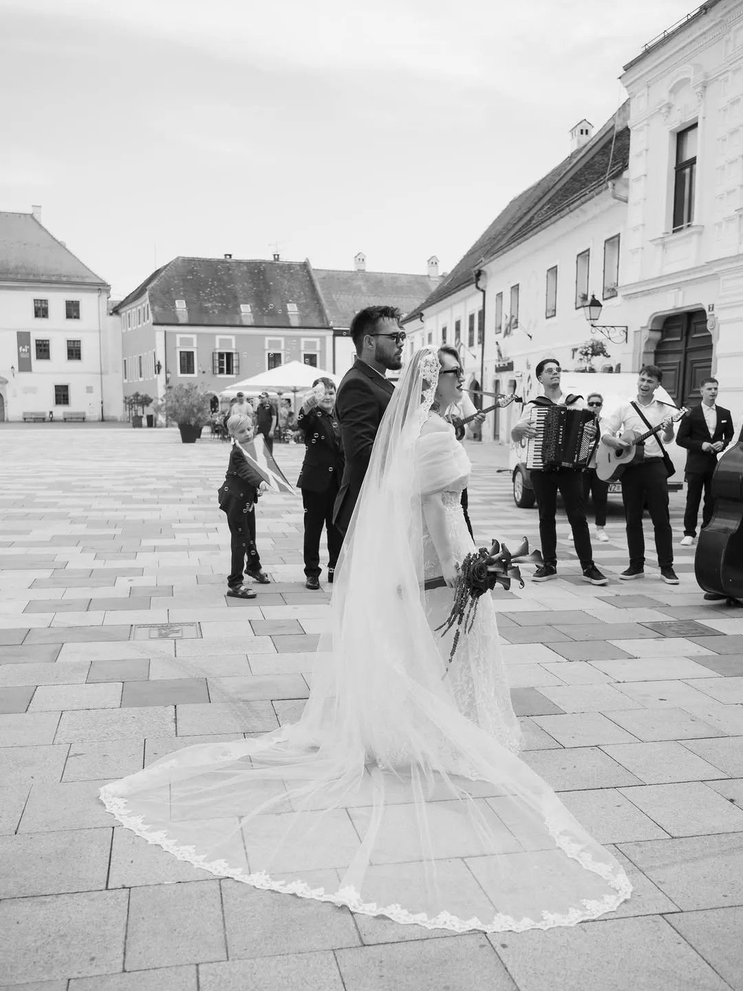 The couple walk across a town square with a long veil while a tamburica band, a traditional Croatian string band, plays behind them
